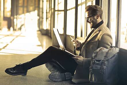 Foto: Studierender sitzt auf dem Boden, hat seinen Laptop auf dem Schoß und blickt in sein Handy