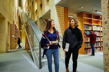 Foto: Zwei Studierende gehen durch die Bibliothek der Universität Münster und unterhalten sich.