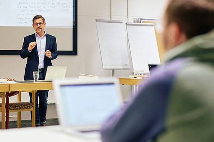 Foto: Ein Dozent steht an einem Tisch in einem Seminarraum und unterrichtet Studierende