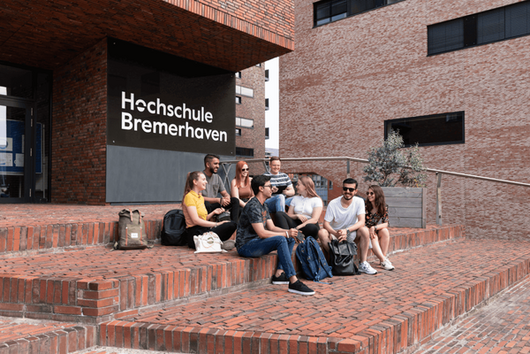 Foto: Studierende sitzen vor dem Eingang zur Hochschule.