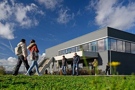 Foto: Blick auf die Mensateria auf dem Campus Nord der Uni Würzburg. Im Vordergrund gehen Studierende über eine grüne Wiese.