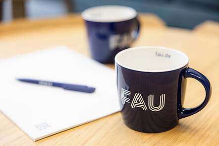 Foto: FAU-gebrandete Tassen, Kugelschreiber und Block auf einem Schreibtisch.