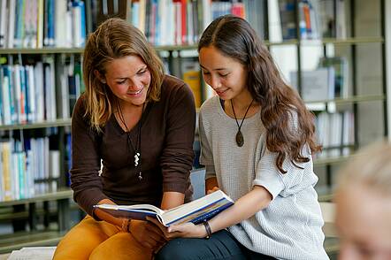 Foto: Zwei Studentinnen sitzen in der Bibliothek und lesen in einem Buch.