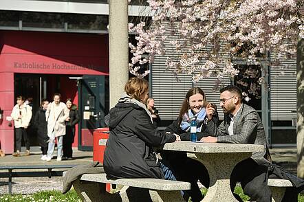 Foto: Drei Studierende sitzen an einem Tisch auf dem Campus der Katholischen Hochschule Mainz und unterhalten sich