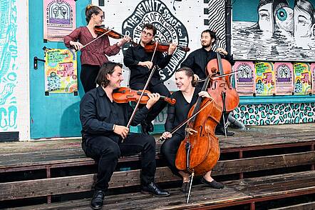 Foto: Fünf Studierende mit Streichinstrumenten sitzen auf einer langen Holzbank und musizieren gemeinsam