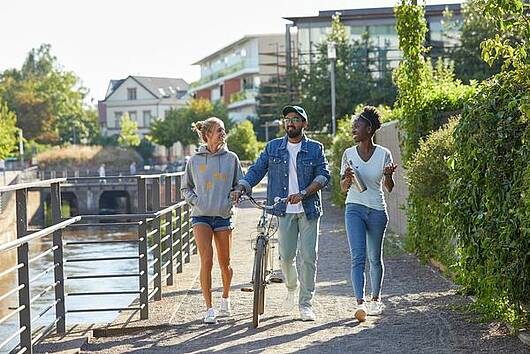 Foto: Studierende der RPTU Kaiserslautern spazieren am Fluss entlang und unterhalten sich.