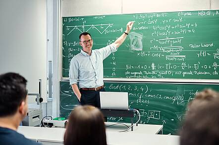 Lehrsituation an der Hochschule Aalen (Foto: Hochschule Aalen/Jan Walford) Foto: Lächelnder Dozent steht an der Tafel, im Vordergrund Köpfe von Studierenden