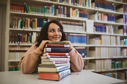 Foto: Studentin sitzt an einem Tisch in der Bibliothek und stützt das Kinn auf einen Stapel Bücher