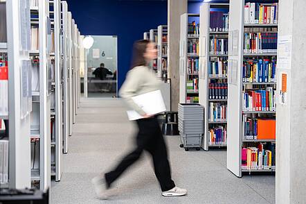 Foto: Eine Studentin geht mit Unterlagen unter dem Arm durch die Gänge der Bibliothek der Hochschule Neu-Ulm