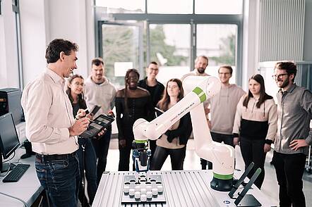 Foto: Ein Dozent steht im Labor und erklärt einer Gruppe Studierender einen Roboterarm