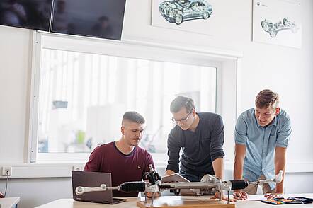 Foto: Drei Studenten sitzen und stehen vor einem technischen Modell und blicken in Laptop und Tablet