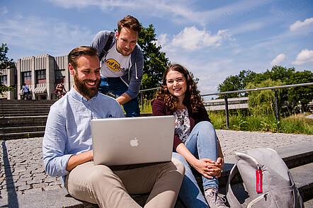 Foto: Eine Studentin uns zwei Studenten sitzen auf einer Treppe auf dem Campus und blicken in einen Laptop