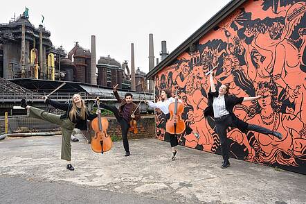 Studierende der HfM Saar vor dem Weltkulturerbe Völklinger Hütte (Foto: Iris M. Maurer) Foto: Studierende halten Instrumente in der Hand und springen vor einer bemalten Mauer in die Luft