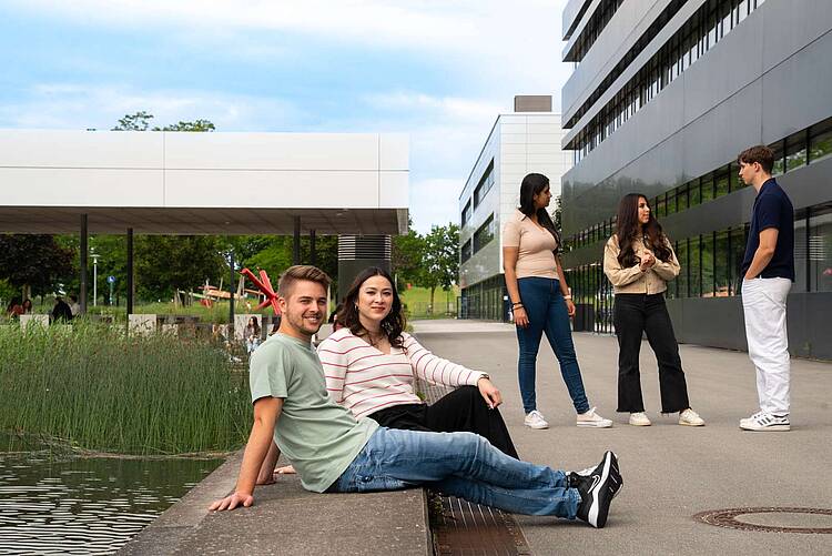 Foto: Zwei Studierende sitzen auf einer Bank auf dem Campus der Hochschule Neu-Ulm, hinter Ihnen Hochschulgebäude und ein kleiner Teich