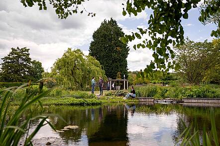 Foto: Blick auf den grünen Campus Haste mit einem See im Vordergrund, an dem sich Studierende aufhalten